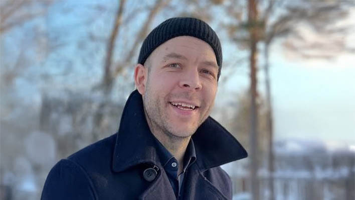 Glad man med sotarmössa och svart jacka , bakom finns det träd